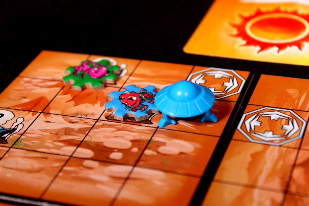 Colorful board game components including a blue saucer, alien tokens, and directional markers on a desert-themed grid board.