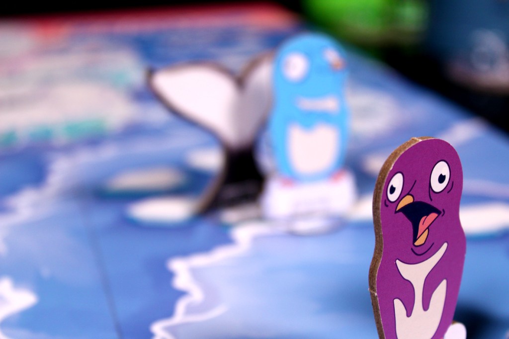 Close-up of a colorful penguin game piece on a playful board with a whale tail in the background.