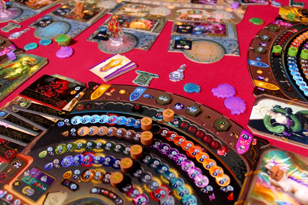 A top-down view of a colorful board game setup featuring various player boards, cards, and game tokens on a red table. The game includes elements like potion ingredients, currency tokens, and unique character pieces.