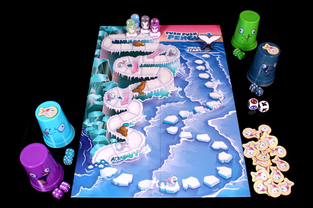 A colorful board game setup featuring the game 'Push Push Penguin' with a game board, penguin cups, dice, and fish tokens arranged on a black surface.