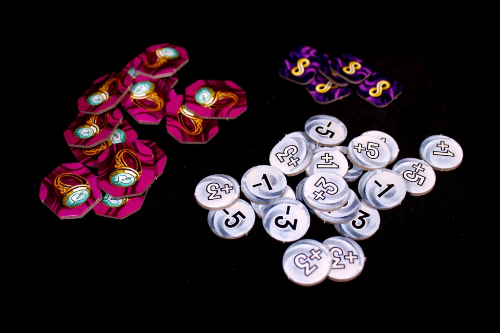 A collection of game tokens featuring various designs and values, including hexagonal tokens with rings and round tokens displaying numbers and symbols, arranged on a black background.