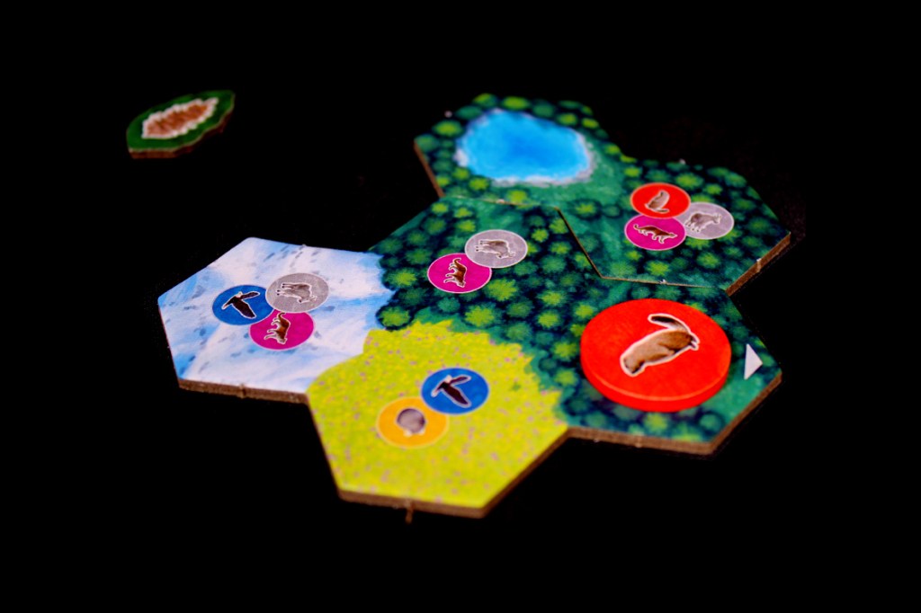 A close-up image of hexagonal game tiles from the board game 'Cascadia: Alpine Lakes,' featuring various wildlife tokens and a blue lake on a lush green background.
