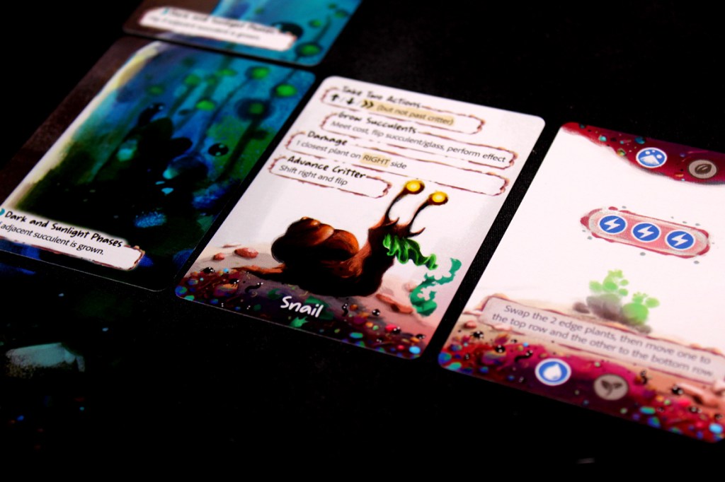 Close-up of game cards from the board game 'Glass Garden', showcasing a snail character and succulent plant actions.