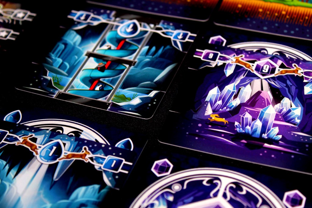 Close-up of game cards from Tranquility: The Descent, featuring colorful illustrations of caves, crystals, and animals, arranged on a dark surface.
