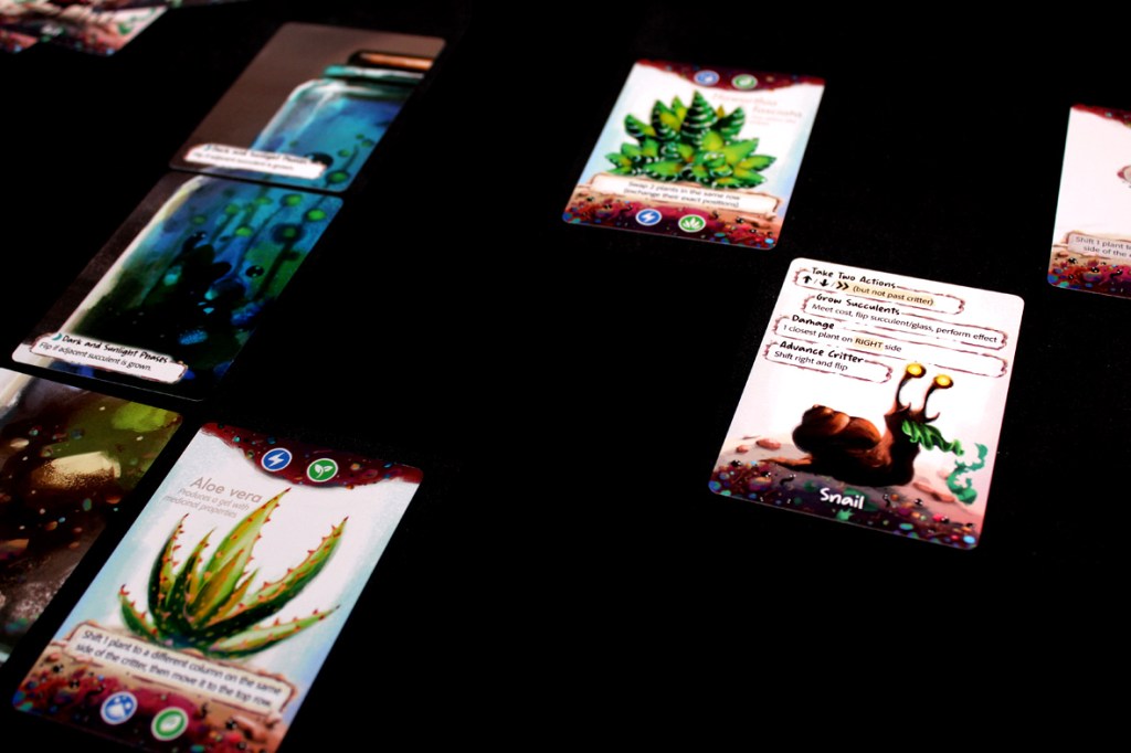 A game setup featuring various cards from the board game Glass Garden, depicting plants and a critter on a dark surface.