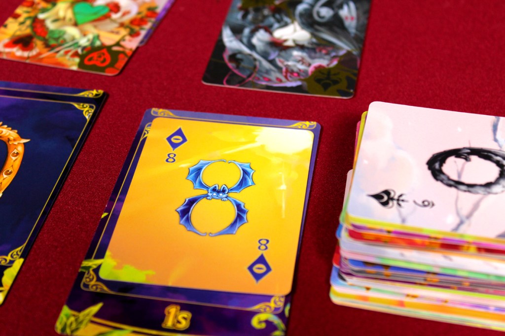 A close-up view of colorful game cards on a red tablecloth, featuring a prominent yellow card with a bat illustration, an 8 of diamonds, and various other artistic cards in the background.