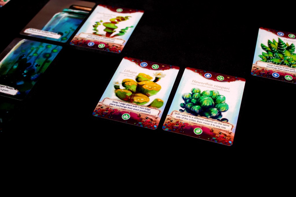 A close-up image of several plant cards from the board game Glass Garden, featuring colorful illustrations of different plants on a dark background.