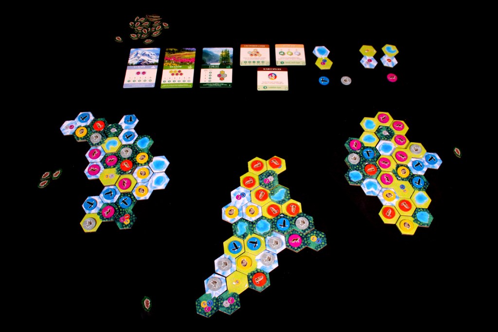 Game board layout of Cascadia: Alpine Lakes, featuring multiple hexagonal tiles in colorful patterns representing different habitats, wildlife tokens, and scoring cards displayed above.