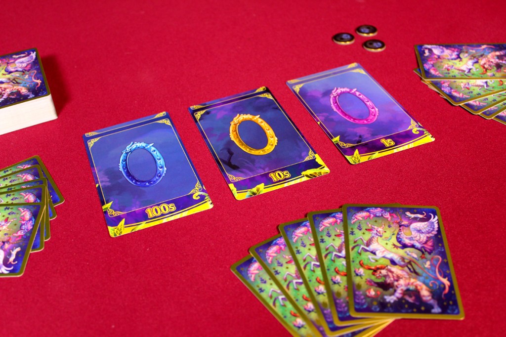 Colorful card game components displayed on a red table, featuring digit cards labeled '100s', '10s', and '1s', alongside additional illustrated cards depicting fantastical creatures.