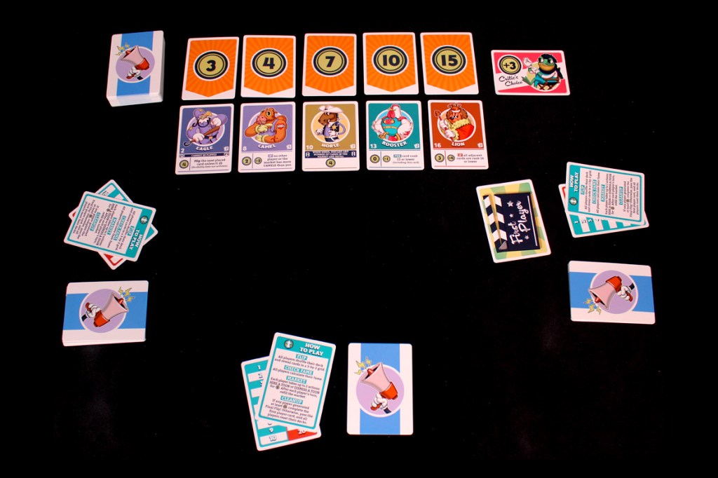 An overhead view of a tabletop game setup for FlipToons, displaying player cards and a market row with various character cards and point markers on a black background.