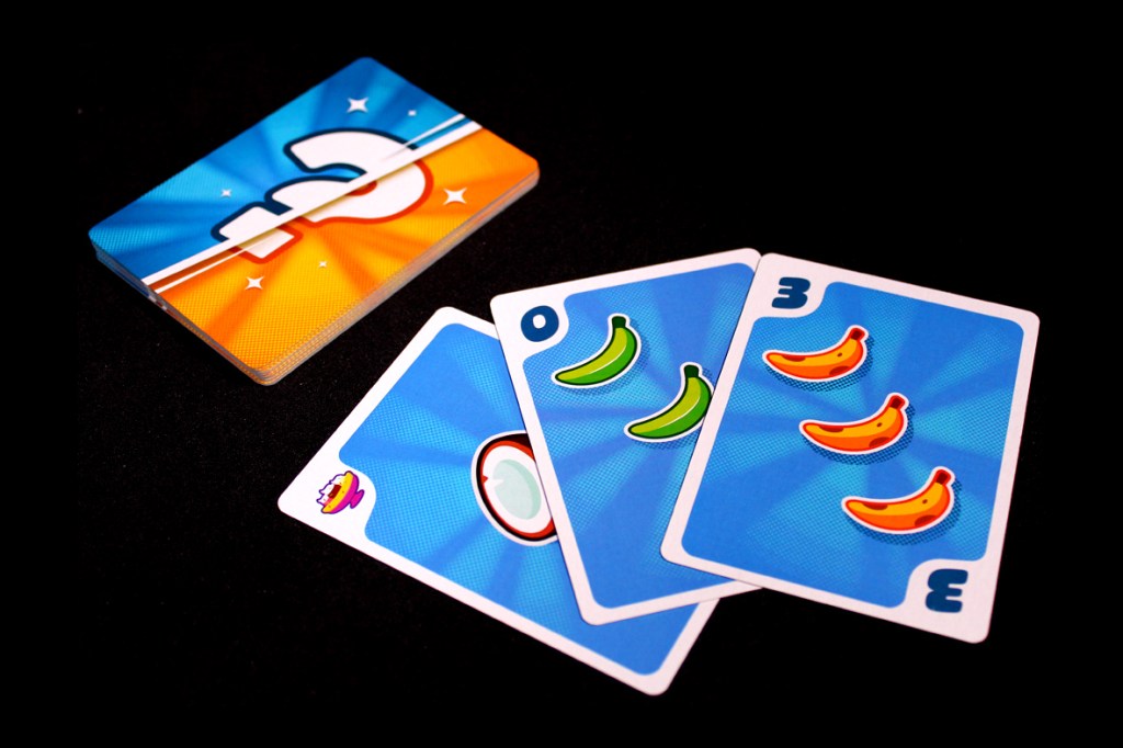 A close-up of several colorful game cards from the board game Fifty Fruity. The cards feature banana and coconut illustrations, along with a central card that displays a number. The background is black, highlighting the vibrant colors of the cards.