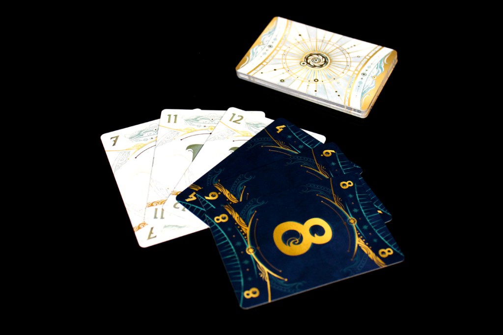 A close-up view of game cards from the board game 'Take Time,' featuring numbered cards (3, 7, 11, 12) in light colors and cards (4, 8) in dark blue with golden designs, alongside a deck box decorated with a clock motif.