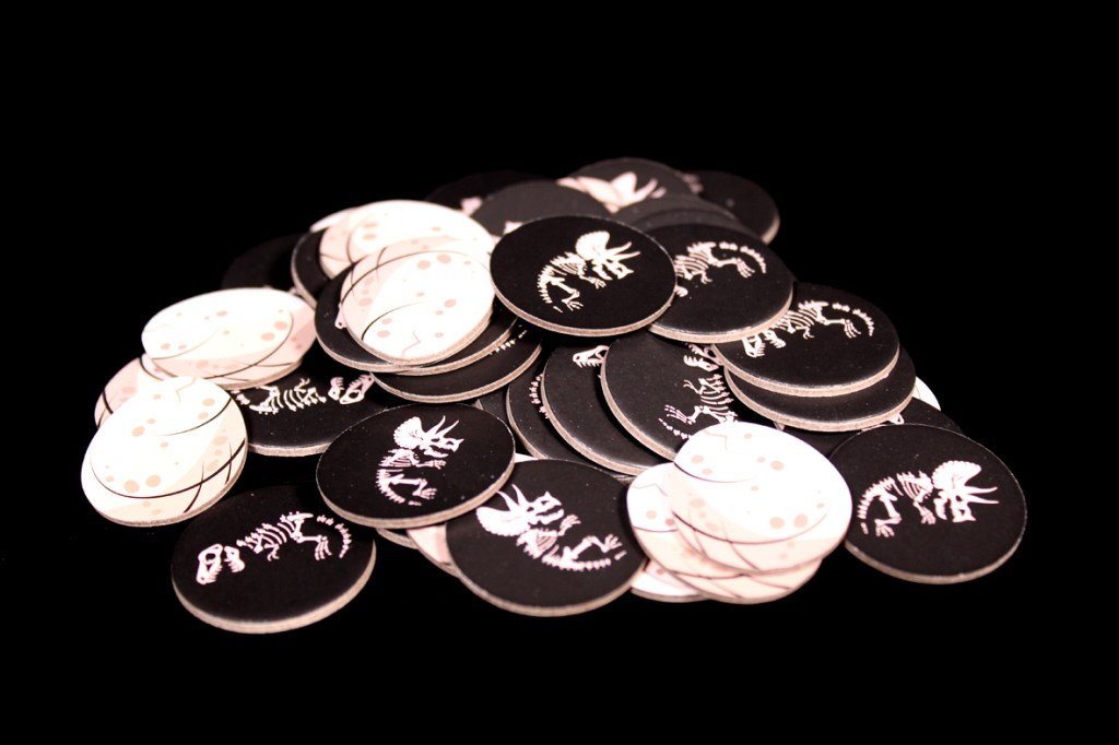 A pile of round game tokens featuring dinosaur skeletons and eggs, presented on a black background.