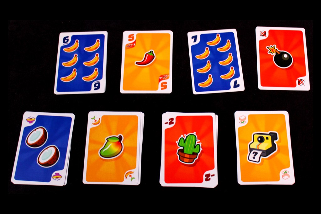 An arrangement of game cards from the board game 'Fifty Fruity', featuring illustrations of bananas, a chili pepper, a bomb, coconuts, a cactus, and a camera, on a black background.