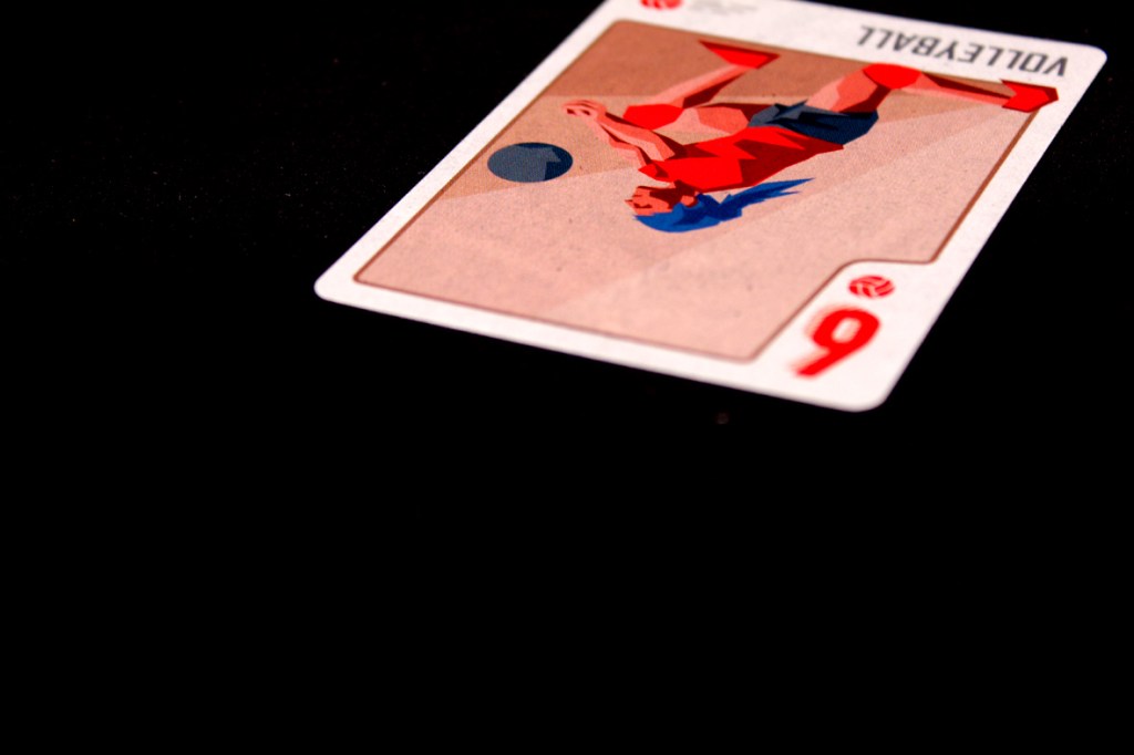 A close-up of a volleyball card featuring a player in red and blue attire, positioned to hit a volleyball. The card is positioned at an angle, showing the number 9 and the word 'VOLLEYBALL' on the top.