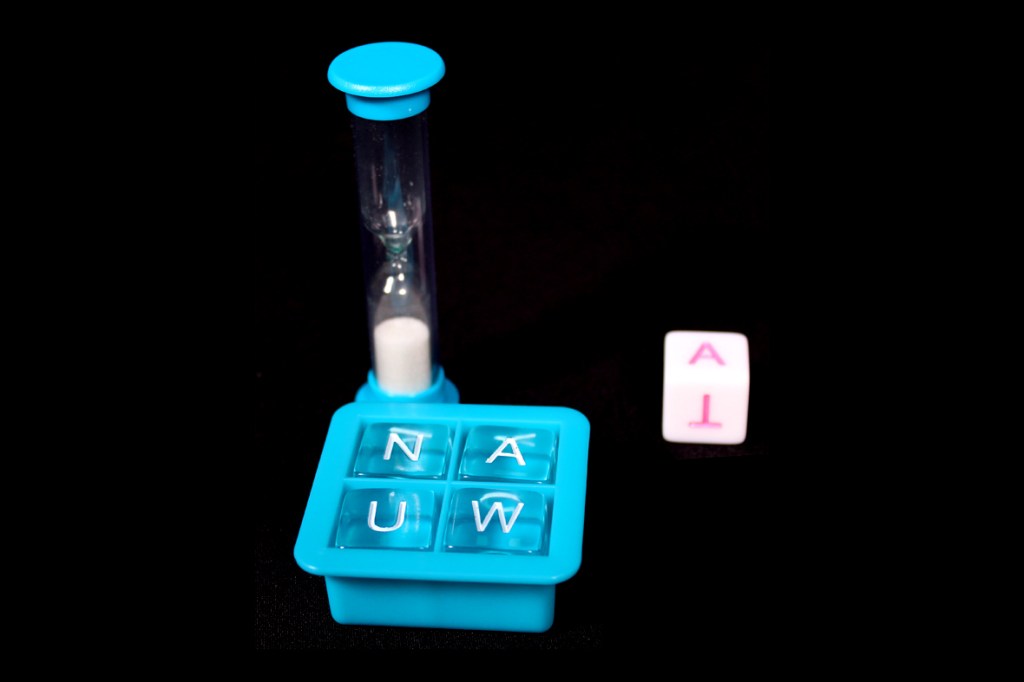 An hourglass timer alongside a blue container with letter tiles displaying 'N', 'A', 'U', and 'W', with a single white die showing 'A' on a black surface.
