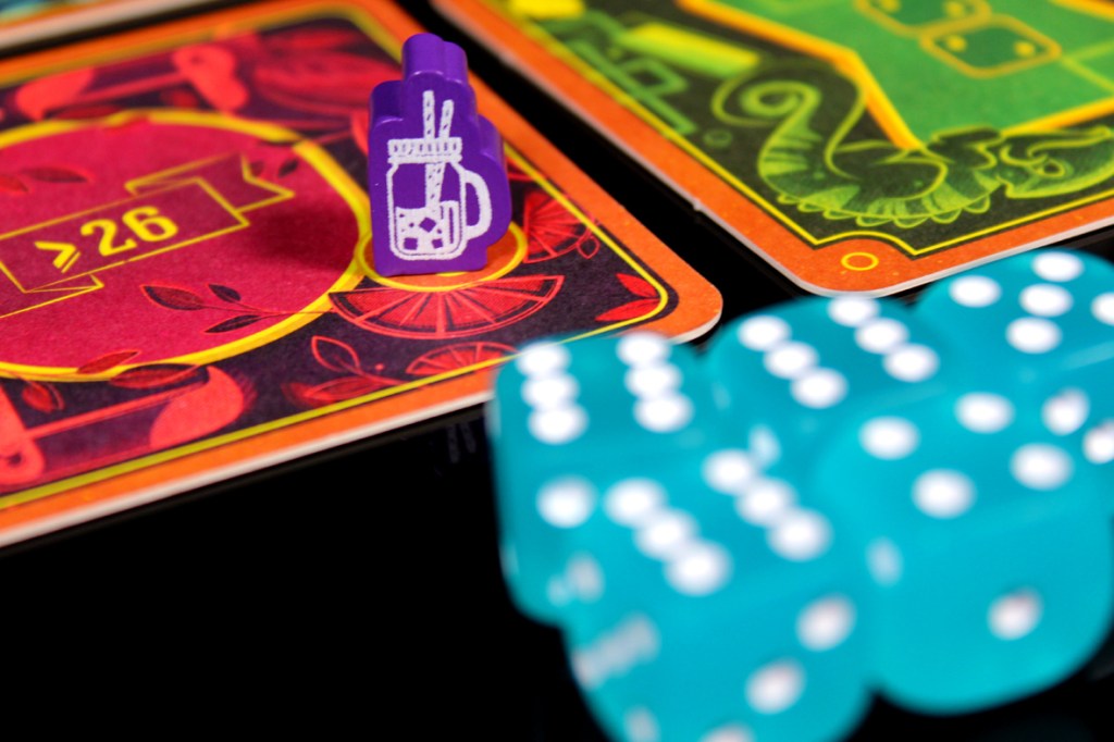 Close-up of game components for Piña Coladice, featuring a purple cocktail token placed on a colorful game board next to teal dice.
