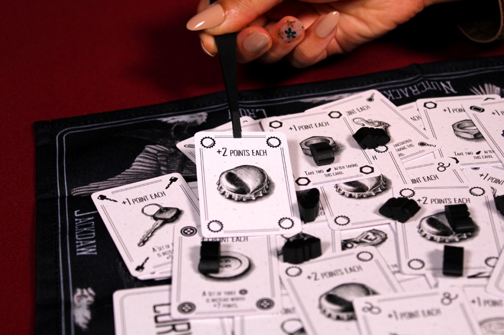A hand holding tweezers is picking up a card from a game mat filled with various game cards and tokens.
