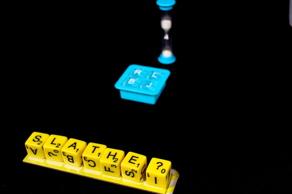 Close-up of yellow letter tiles showing the word 'SLATHE?' on a scoring board, with an hourglass timer and a blue ice tray in the background.