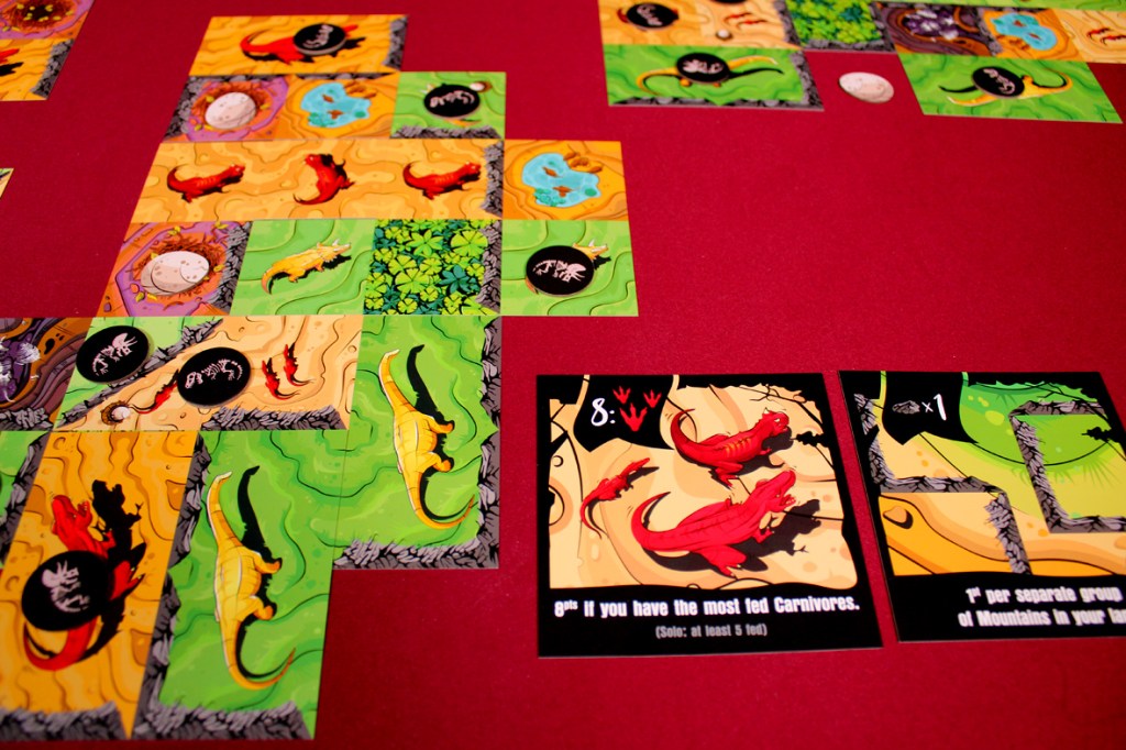 A colorful board game setup featuring various dinosaur-themed cards and tiles on a red table. The game includes goal cards for scoring and tiles depicting dinosaurs, eggs, and terrain.