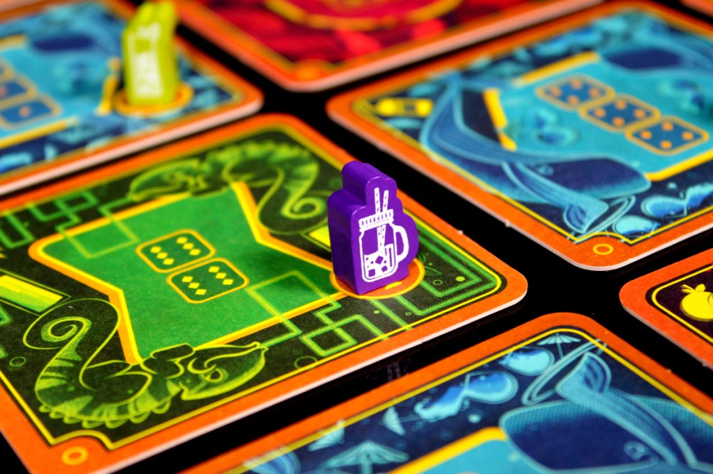 A close-up view of a colorful board game featuring various coasters and game pieces, including a purple token shaped like a drink mug placed on a green coaster with dice illustrations.