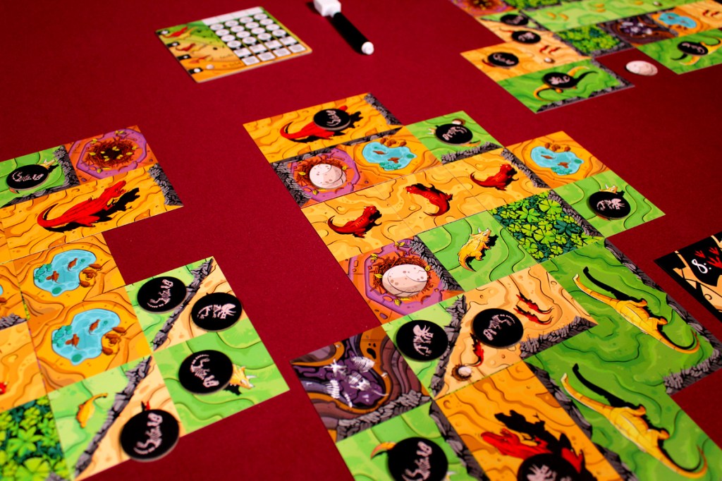 A tabletop board game scene featuring colorful tiles with dinosaur illustrations, set on a maroon tablecloth. The game includes various terrain types and tokens, indicating gameplay in progress.