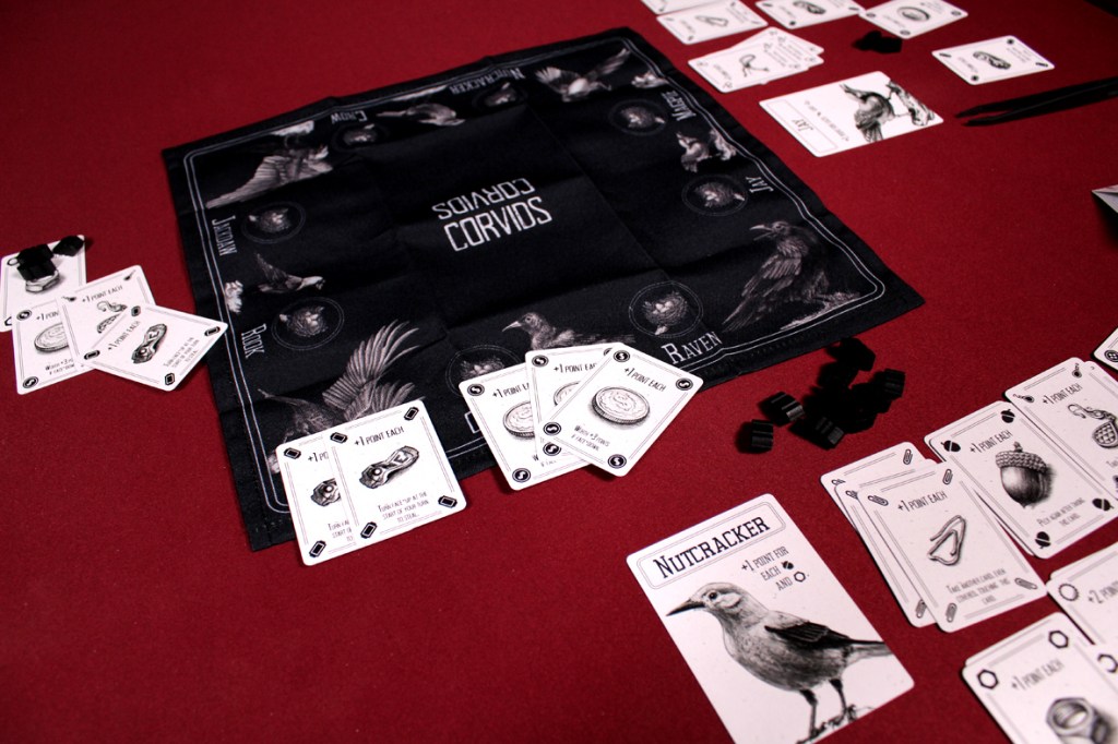 A tabletop game setup for Corvids, featuring a black game mat with illustrations of crows, laid out cards, feather tokens, and tweezers on a red surface.