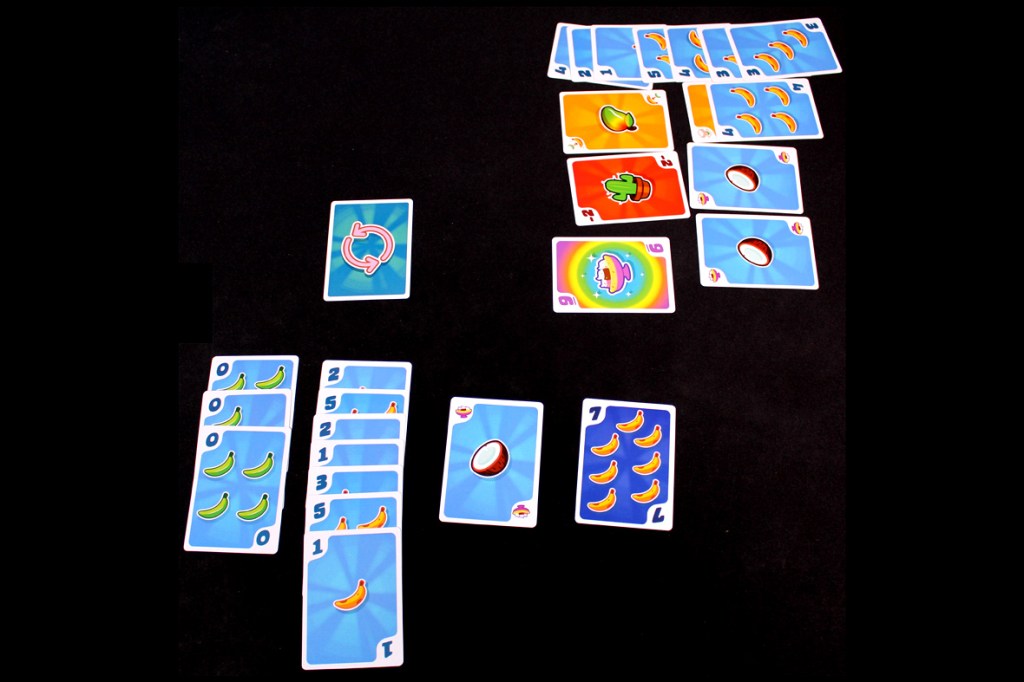 An overhead view of a tabletop game setup for Fifty Fruity, featuring various colorful cards laid out, including banana and coconut illustrations. The Turn Direction Card is prominently displayed in the center.