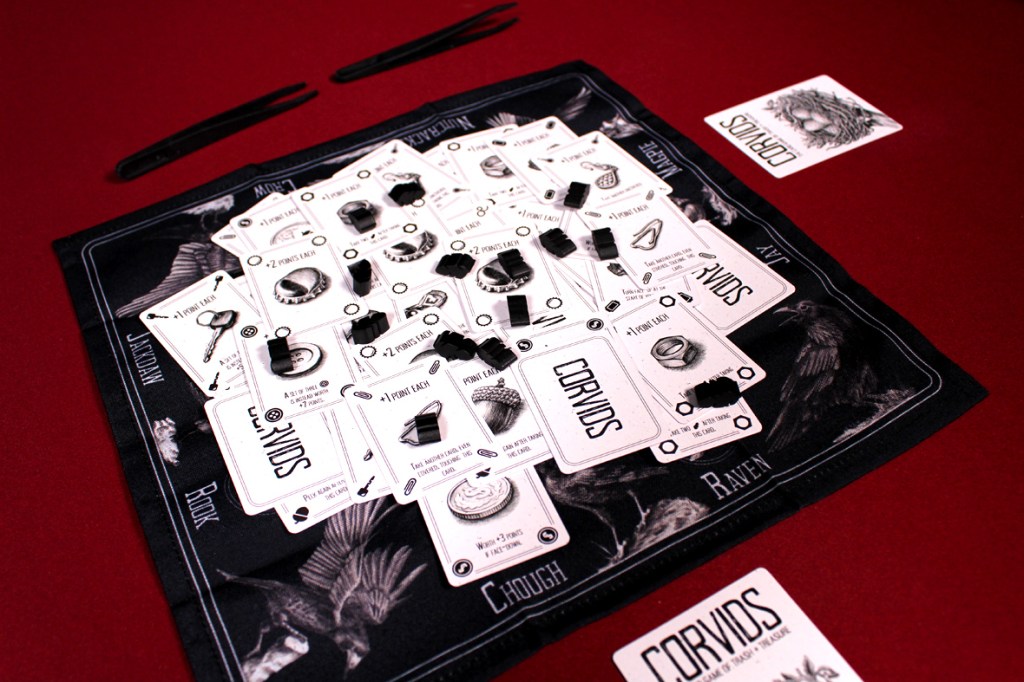 A game setup for Corvids featuring a black and white game mat with various cards and trinket tokens scattered on top.