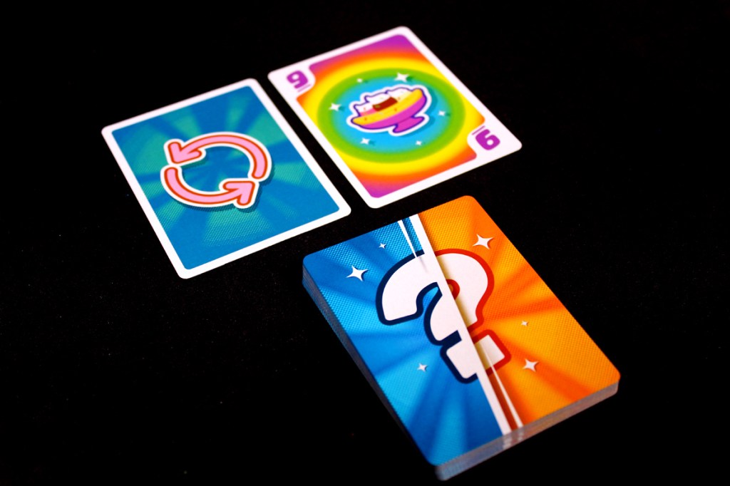 Two playing cards on a black surface: a blue card with a circular arrow symbol and a colorful card labeled with numbers '9' and '6', next to a deck with a question mark design.
