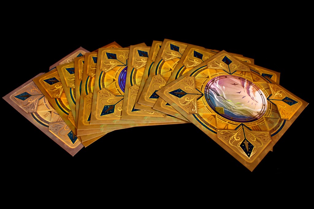 A stack of intricate game cards featuring ornate gold borders and colorful illustrations, displayed against a black background.