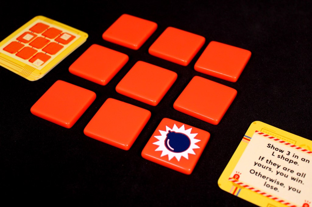 A close-up view of a game setup for Bombastic, featuring a 3x3 grid of red tiles and two action cards with instructions.