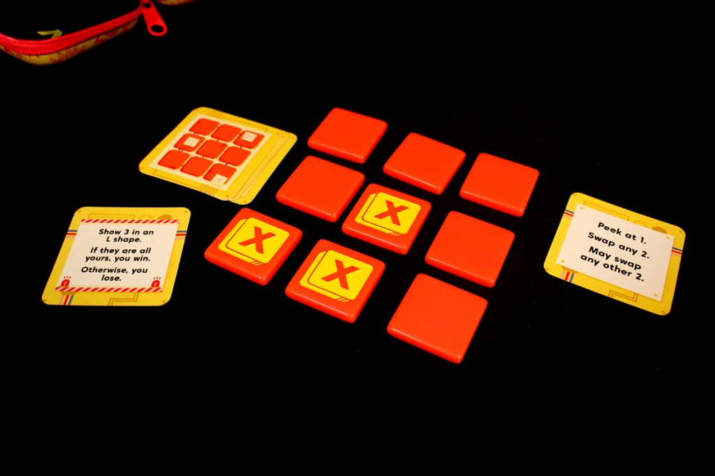 A close-up of game components for Bombastic, featuring red game tiles, action cards with instructions, and a colorful carrying case.