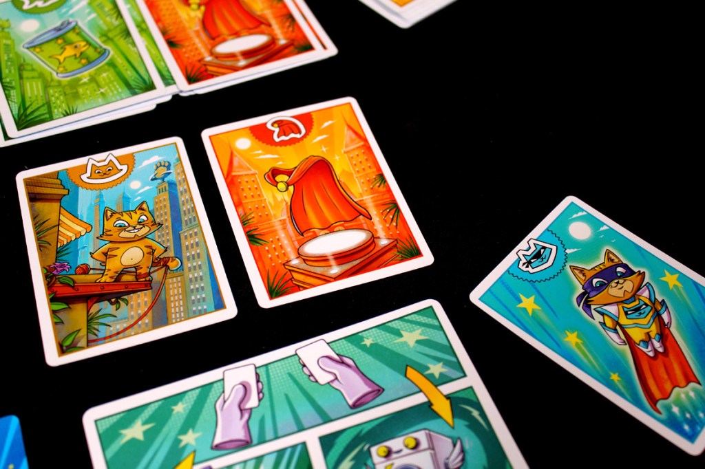 A colorful tabletop scene featuring playing cards from the game Super Meow, including images of a cat, a cape, and various game mechanics illustrated with bright designs.