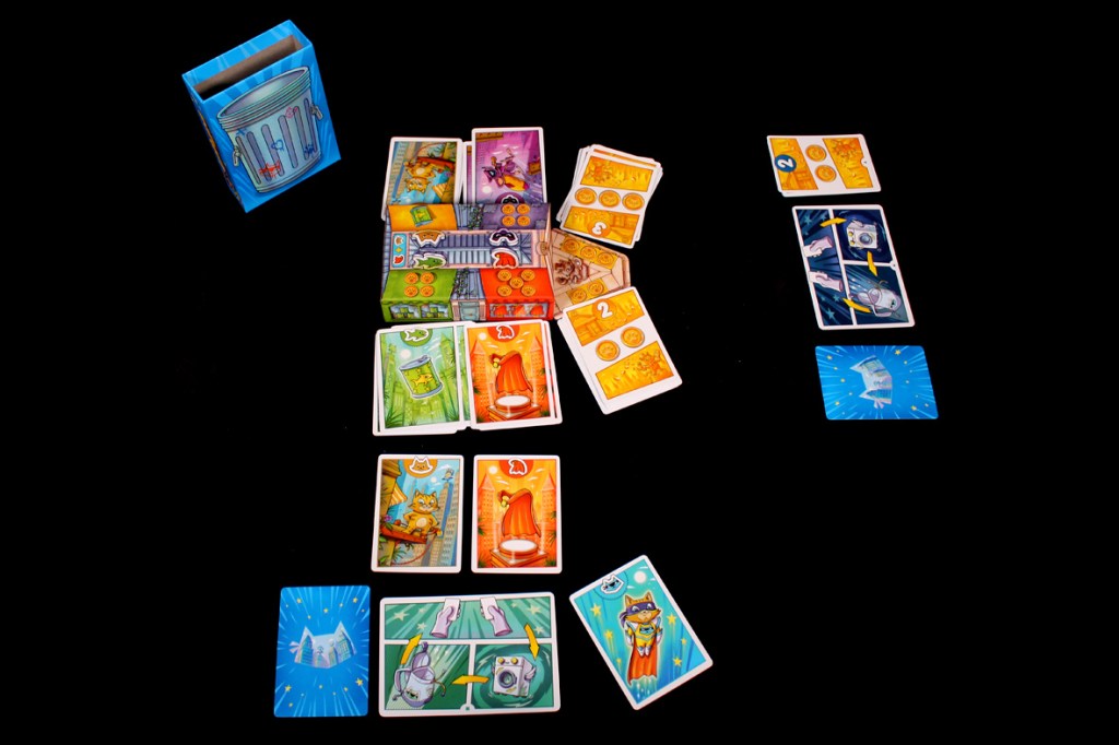 A colorful setup for the card game 'Super Meow', featuring a trash box, a market area with character cards, and various game components laid out on a black background.