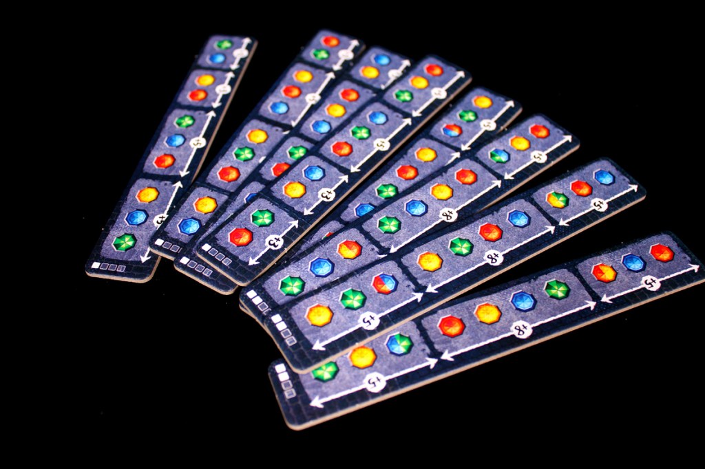 A fan of scoring tiles from the board game Umbrella arranged in a scattered layout, displaying colorful patterns with gems in red, blue, green, and yellow on a dark background.