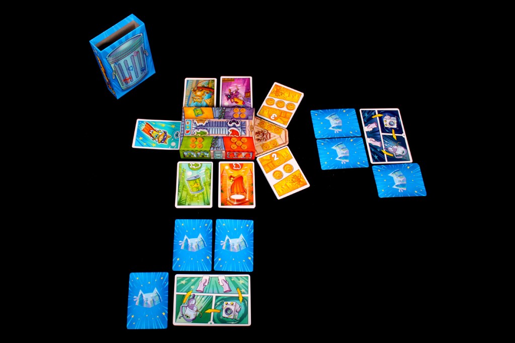 A tabletop layout for the game Super Meow, featuring a colorful setup including character and action cards, a market area with coins, and a box for storing game components.