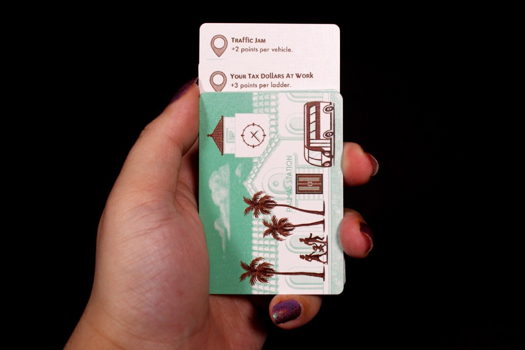 A hand holding a game card with two point scoring instructions: 'Traffic Jam' and 'Your Tax Dollars At Work', featuring illustrated palm trees and a bus.