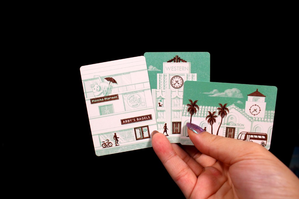 A hand holding three illustrated cards featuring local businesses and landmarks, including 'Matcha Marmot,' 'Abby's Bagels,' and 'Palm Station.' The cards are designed with a teal color scheme and depict a person walking and a bicycle.