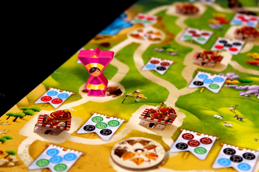 A close-up view of a colorful board game featuring a pink player token, various resource cards, and a scenic game board with pathways and settlements.