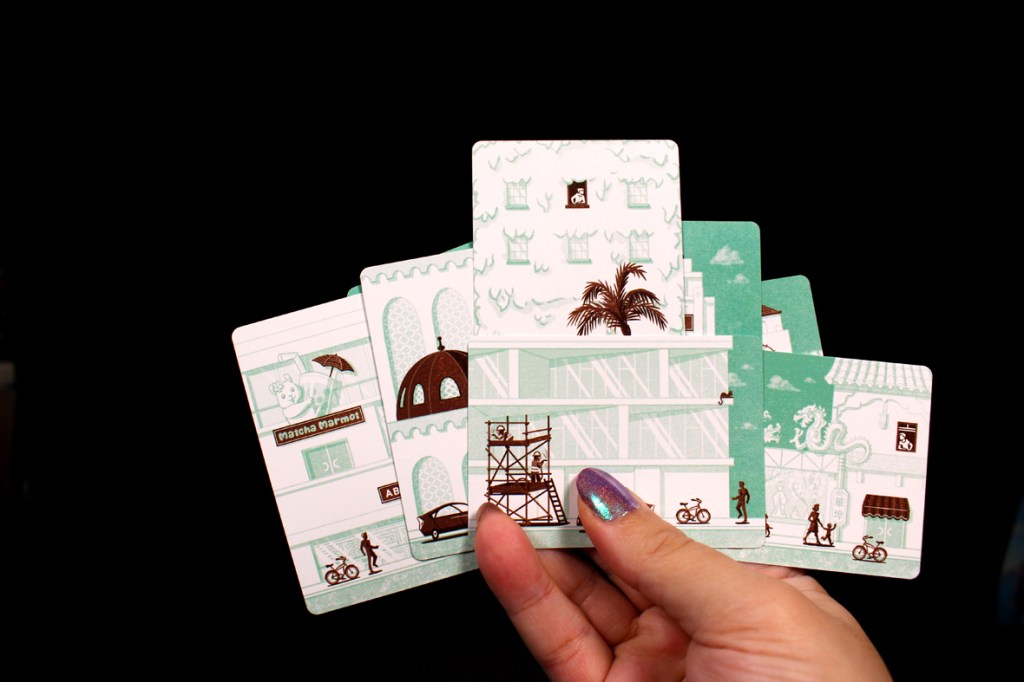 A hand holding several illustrated cards depicting buildings, cyclists, and palm trees, with a dark background.
