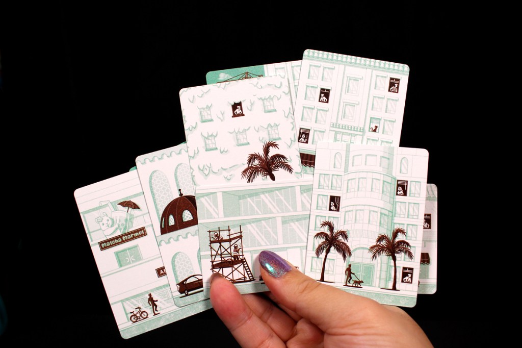 A hand holding several illustrated cards featuring urban scenes, including buildings, palm trees, bicycles, and various characters engaged in activities.