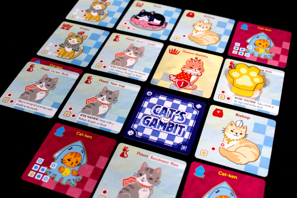 A flat lay of the game Cat's Gambit: Queen Aurelia, featuring various character and action cards with illustrations of cats, paw prints, and a colorful game board design.