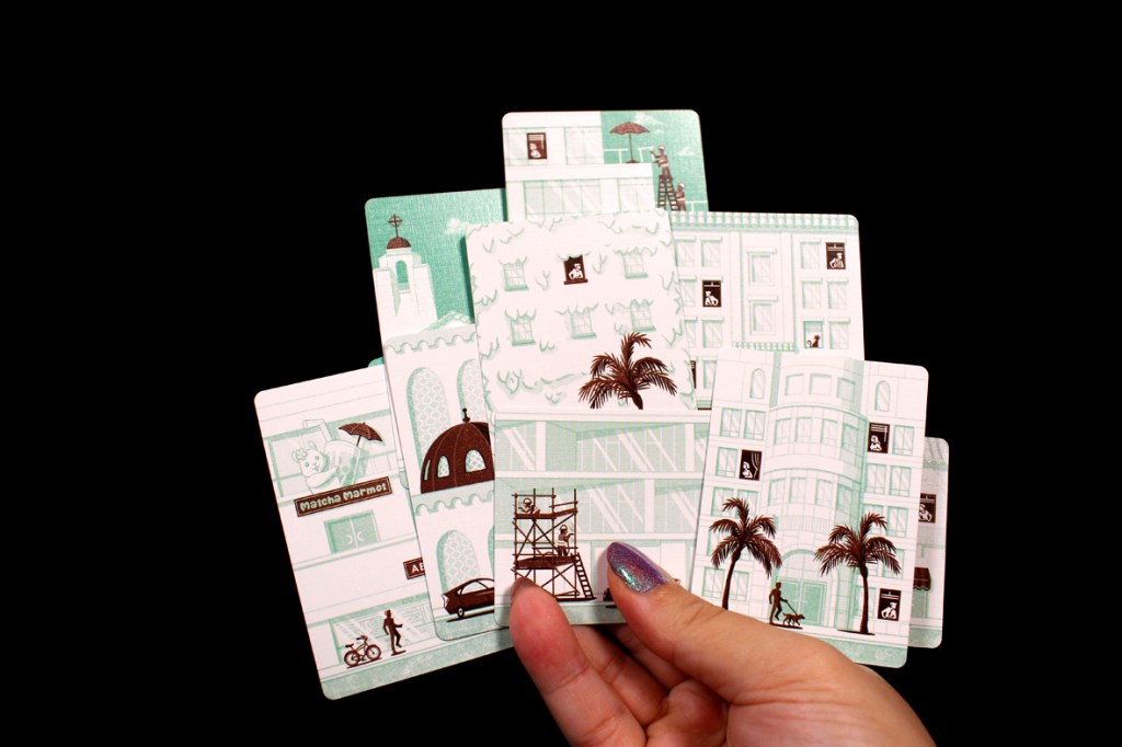 A hand holding several illustrated cards featuring stylized buildings, palm trees, and urban scenes against a black background.
