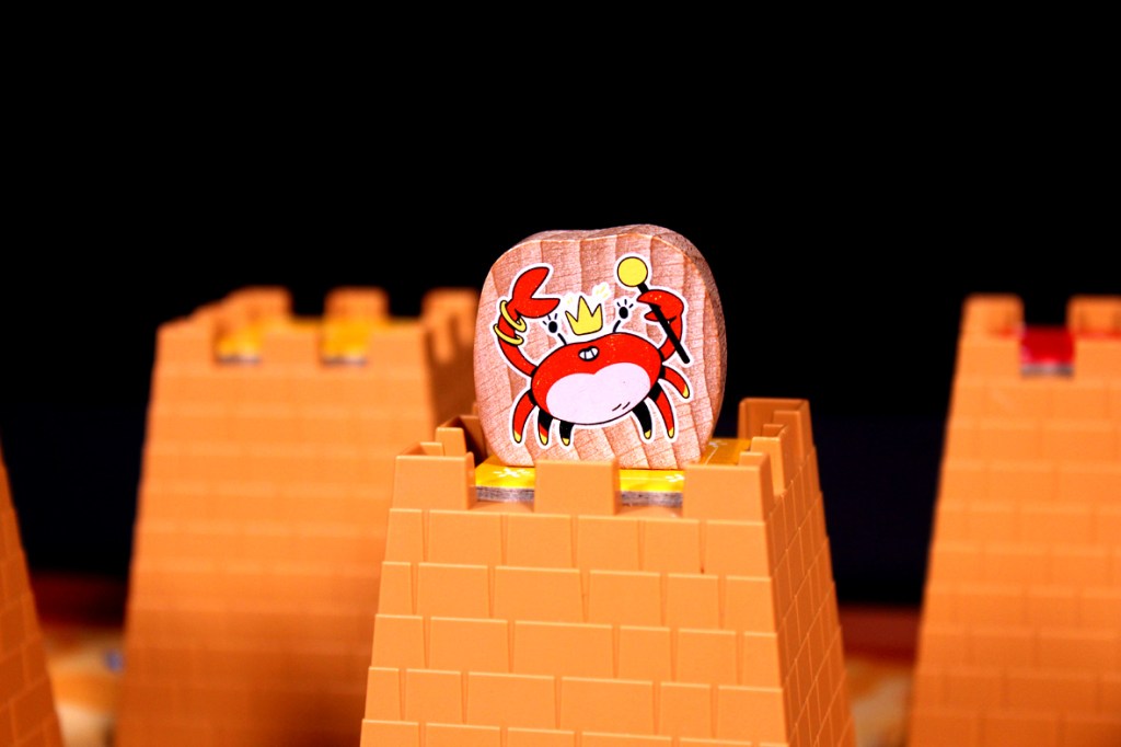 A wooden game piece depicting a cartoon crab with a crown, positioned on top of an orange block structure.