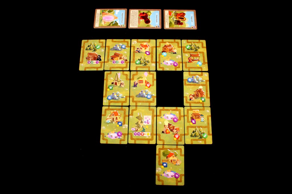A layout of cards for the board game 'A Dragon's Gift', depicting various village buildings and resources arranged in a specific pattern.
