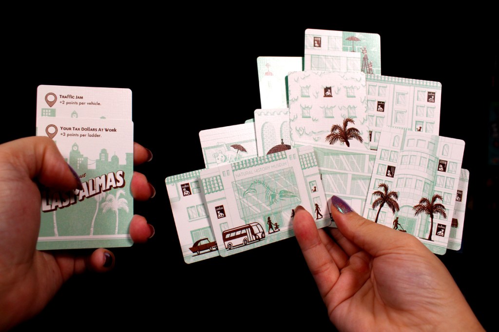 A person's hand holding game cards featuring illustrations of buildings, vehicles, and tropical palm trees, with text descriptions for game mechanics like 'Traffic Jam' and 'Your Tax Dollars At Work.'