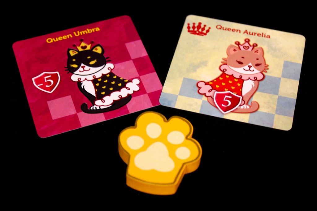 Two character cards featuring Queen Umbra and Queen Aurelia from the game Cat's Gambit, with a yellow paw print token in the foreground.