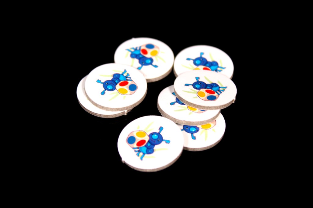 A collection of circular game tokens with colorful insect designs, featuring blue insects with red, yellow, and blue spots, placed against a black background.