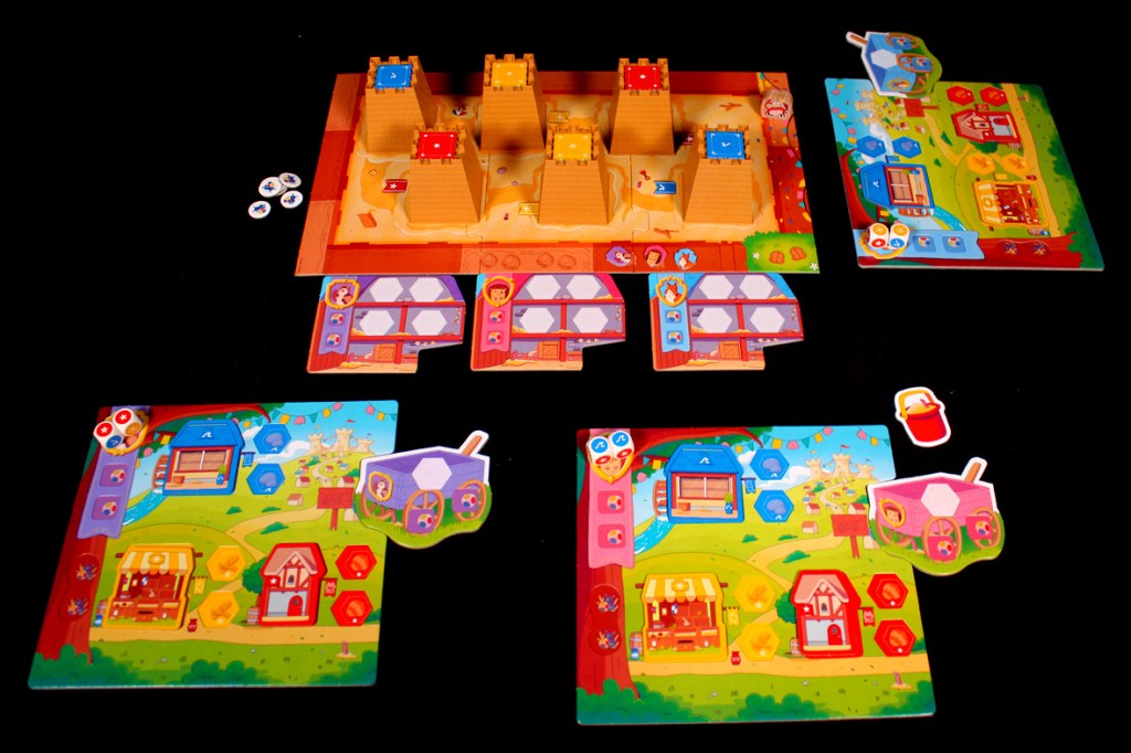 A colorful tabletop board game setup featuring various game boards, tokens, and player pieces arranged on a black surface, with sandy castle structures as the central focus.
